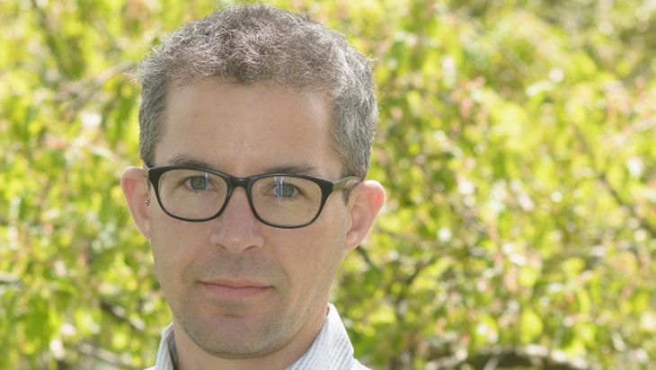 Photo of Alex Meyers wearing a dress shirt and glasses in front of trees