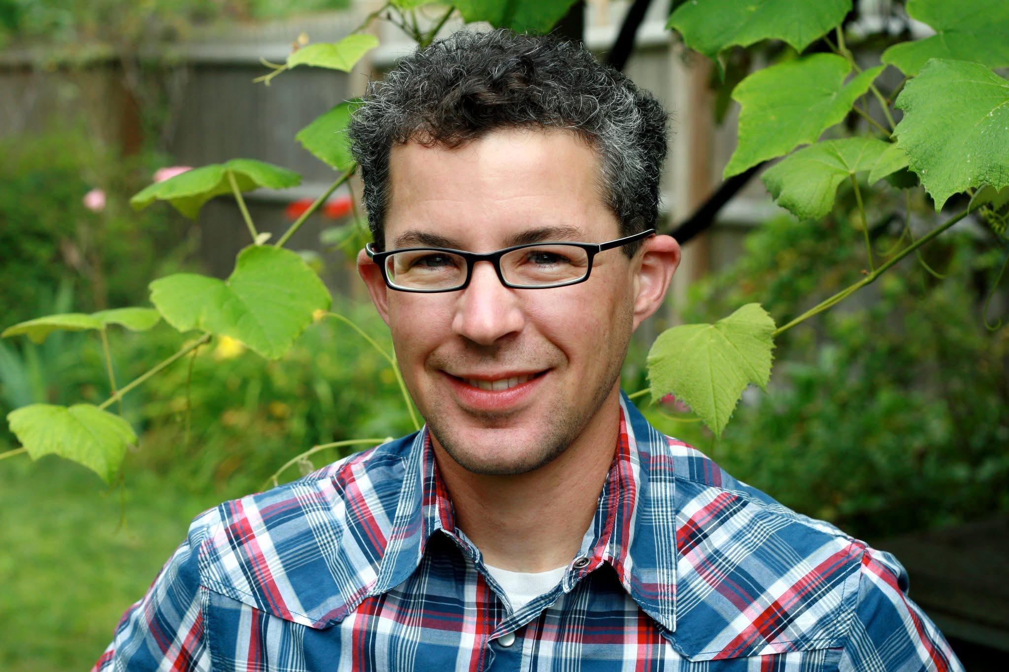 Candid photo of Author Alex Myers in a blue & red plaid shirt with leaves in the background
