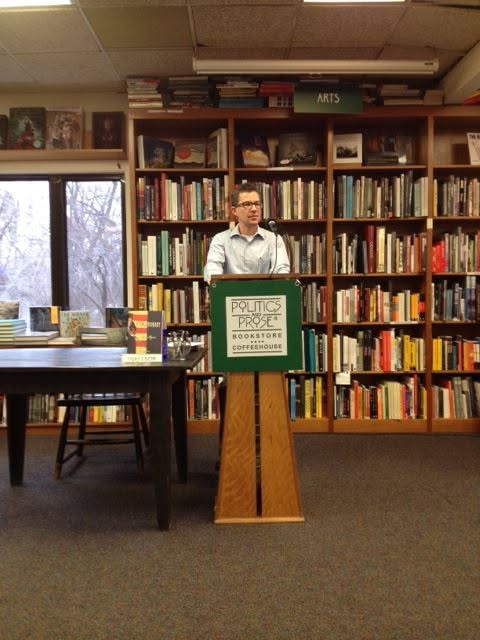Alex at a podium in a library. The podium reads Politics and Prose.