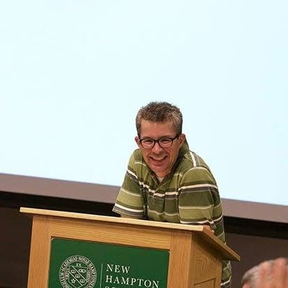Alex giving a lecture in front of a white screen and a podium with New Hampton School on it.